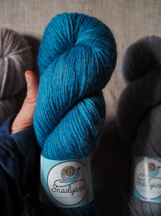Blue skein of yarn held by a hand with 'Snailyarn' label, against a neutral background.