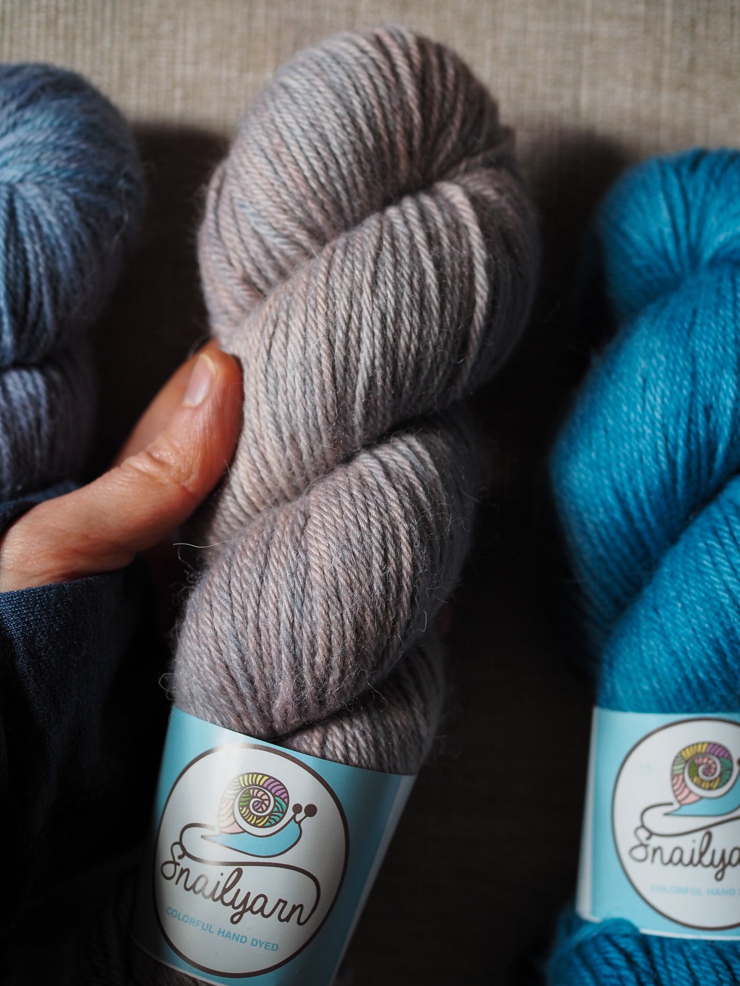 Three skeins of yarn in gray, blue, and turquoise held by a hand on a textured surface.