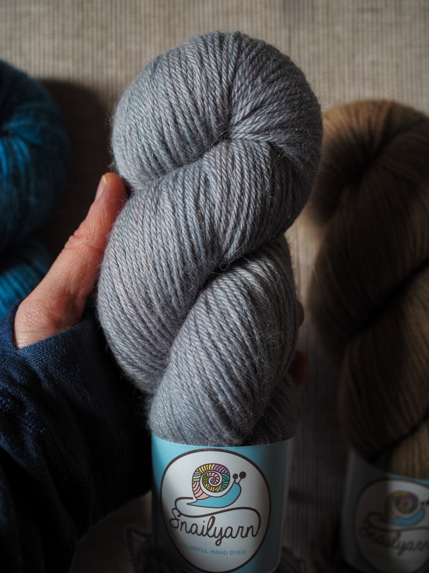 Gray skein of yarn held by a hand with a visible label, against a neutral background.